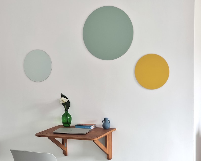 May include: Three round wall decorations in a minimalist home office setting. The largest circle is a light green colour, the medium circle is a light blue colour, and the smallest circle is a yellow colour. A small wooden folding desk with a laptop, a vase with a white flower, and a blue ceramic vase is in the foreground.