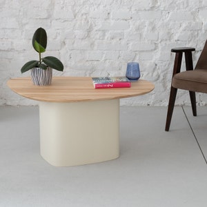 Peut inclure: Une table basse ovale en bois clair avec un piétement blanc. Une petite plante en pot et un livre sont posés sur la table. Un verre bleu est posé sur la table à côté du livre.