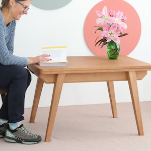 May include: A light-coloured wooden coffee table with angled legs. A person is seated next to the table, holding a book. A vase of pink lilies sits on the table. The table is approximately 76 cm long.