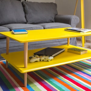 Può includere: Un tavolino da caffè giallo a due livelli con gambe in legno. Il tavolino è su un tappeto a strisce colorate davanti a un divano grigio.