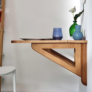 May include: A wooden wall-mounted fold-down desk with a light wood finish. The desk has a triangular support structure and is shown with a blue glass, a green vase with white flowers, and a blue vase.