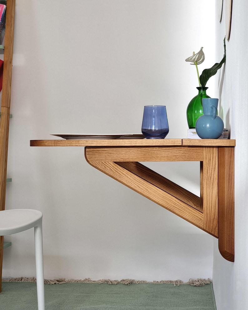 May include: A wooden wall-mounted fold-down desk with a light wood finish. The desk has a triangular support structure and is shown with a blue glass, a green vase with white flowers, and a blue vase.