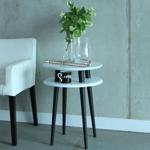 Puede incluir: Mesa auxiliar blanca de dos niveles con patas negras. La mesa es redonda y tiene un jarr&oacute;n blanco con flores blancas en el nivel superior.