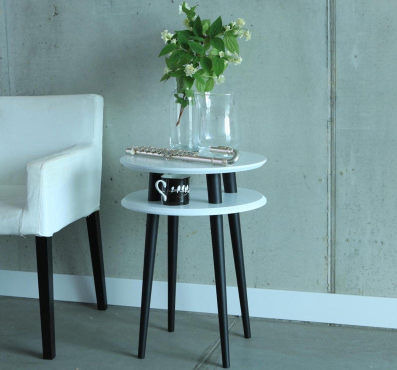 Puede incluir: Mesa auxiliar blanca de dos niveles con patas negras. La mesa es redonda y tiene un jarr&oacute;n blanco con flores blancas en el nivel superior.