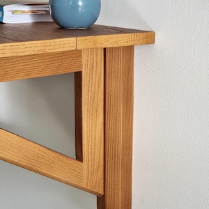 May include: A wooden wall-mounted shelf with a light brown finish. The shelf has a simple design with a triangular support structure and a rectangular top. The shelf is made of solid wood and has a natural grain pattern.