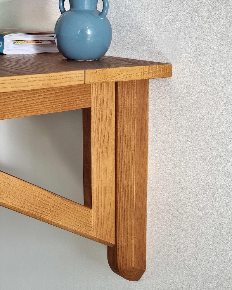 May include: A wooden wall-mounted shelf with a light brown finish. The shelf has a simple design with a triangular support structure and a rectangular top. The shelf is made of solid wood and has a natural grain pattern.