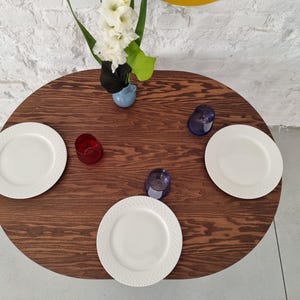 May include: A dark brown oval wooden table with three white plates and three glasses. Two of the glasses are blue and one is red. A vase with white flowers is in the center of the table.
