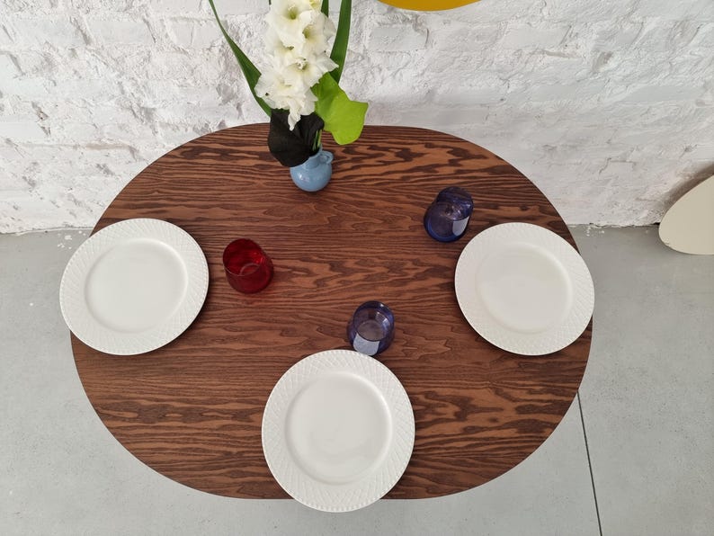 May include: A dark brown oval wooden table with three white plates and three glasses. Two of the glasses are blue and one is red. A vase with white flowers is in the center of the table.
