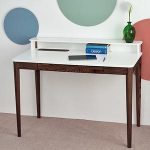 Könnte beinhalten: Ein moderner weißer Schreibtisch mit dunkelbraunen Holzbeinen und einer passenden Schublade. Der Schreibtisch hat eine weiße Oberseite mit einem eingebauten Regal und eine grüne Glasvase mit einer weißen Blume. Zwei blaue Bücher mit dem Wort "Design" stehen auf dem Regal.