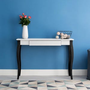 Pode incluir: Uma mesa consola branca com uma gaveta única e quatro pernas pretas curvas. Sobre a mesa, encontra-se um vaso branco texturizado com rosas cor-de-rosa e um cesto de arame preto preenchido com várias esferas decorativas claras.