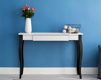 May include: A white console table with a single drawer and four curved black legs. On the table sits a white textured vase containing pink roses and a black wire basket filled with several light-colored decorative spheres.