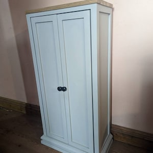 May include: A tall, light grey cabinet with two doors and a wood top. The cabinet has a simple design with a slightly rounded base.