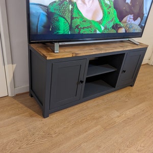 May include: A dark grey TV stand with two doors and a shelf. The stand has a wooden top and is sitting on a light wood floor.