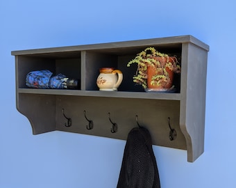 Könnte beinhalten: Ein dunkelbraunes, hölzernes Wandregal mit zwei offenen Fächern und fünf schwarzen Metallhaken. Das Regal hat eine dekorative, abgerundete Kante und ist an einer hellblauen Wand montiert. Ein blaues und weiß gemustertes Regenschirm, eine braune Keramiktasse, eine Topfpflanze und ein schwarzer Pullover sind auf dem Regal ausgestellt.