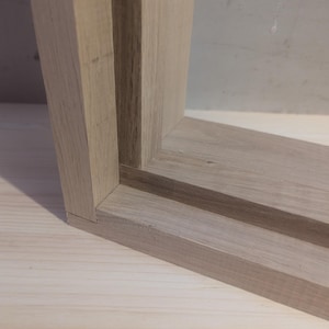 May include: Close-up of a corner of a light-coloured wooden frame. The frame is constructed with precise angles and joints, showcasing the natural grain of the wood. The frame appears to be unfinished, with a smooth, unpainted surface. The wood has a warm, neutral tone.