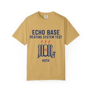 May include: A mustard yellow short-sleeve t-shirt featuring a blue graphic of a radiator with a snowflake icon and red heat waves. The text reads ECHO BASE HEATING SYSTEM TEST and HOTH in blue capital letters centered on the front of the shirt.