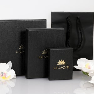 May include: Four black gift boxes with gold lettering. The largest box says "Lilyom" and the smaller boxes say "Lilyom". The boxes are on a white surface with white flowers.