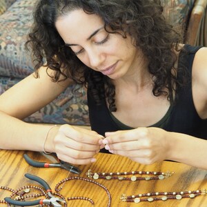 May include: A person is making jewelry with brown and gold beads. They are using pliers to help them create the jewelry. The jewelry is on a wooden table.