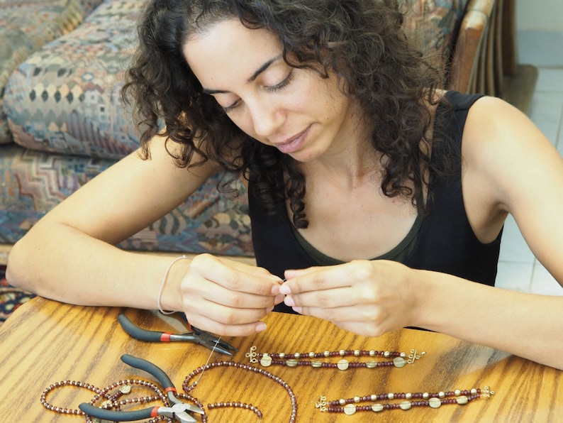 May include: A person is making jewelry with brown and gold beads. They are using pliers to help them create the jewelry. The jewelry is on a wooden table.