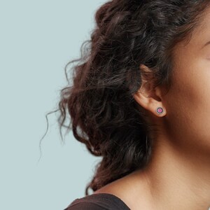 May include: A close-up of a person's ear with a small pink gemstone earring. The earring has a silver setting with a black border.