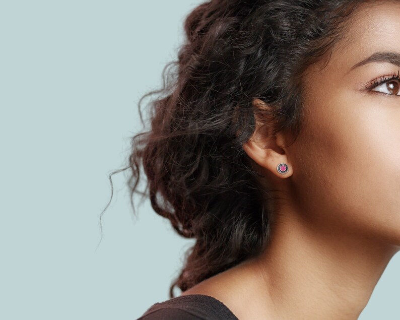 May include: A close-up of a person's ear with a small pink gemstone earring. The earring has a silver setting with a black border.