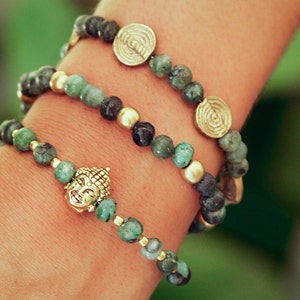 Puede incluir: Tres pulseras superpuestas con cuentas verdes y negras y detalles dorados. La pulsera inferior presenta un dije de cabeza de Buda dorado.