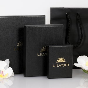 May include: Three black gift boxes with gold lettering, the smallest box says "Lilyom", the middle box says "Lily", and the largest box says "Lil", all boxes are on a white surface with white flowers.