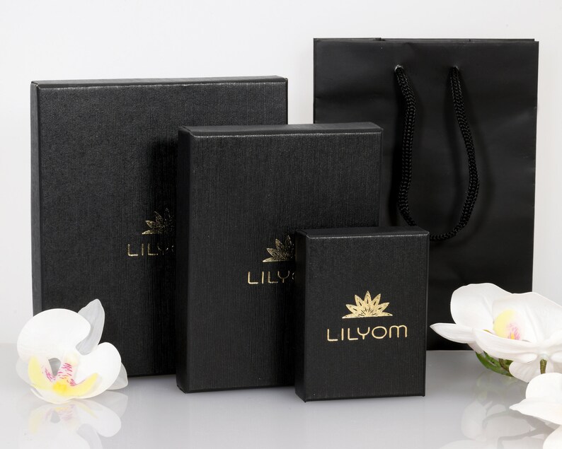May include: Three black gift boxes with gold lettering, the smallest box says "Lilyom", the middle box says "Lily", and the largest box says "Lil", all boxes are on a white surface with white flowers.