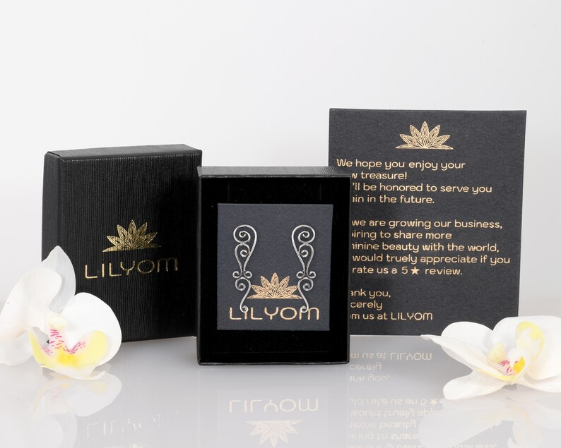 May include: A pair of silver earrings with a delicate, swirling design. The earrings are packaged in a black box with a gold logo that reads "LILYOM". The box is sitting on a white surface with a white orchid flower.