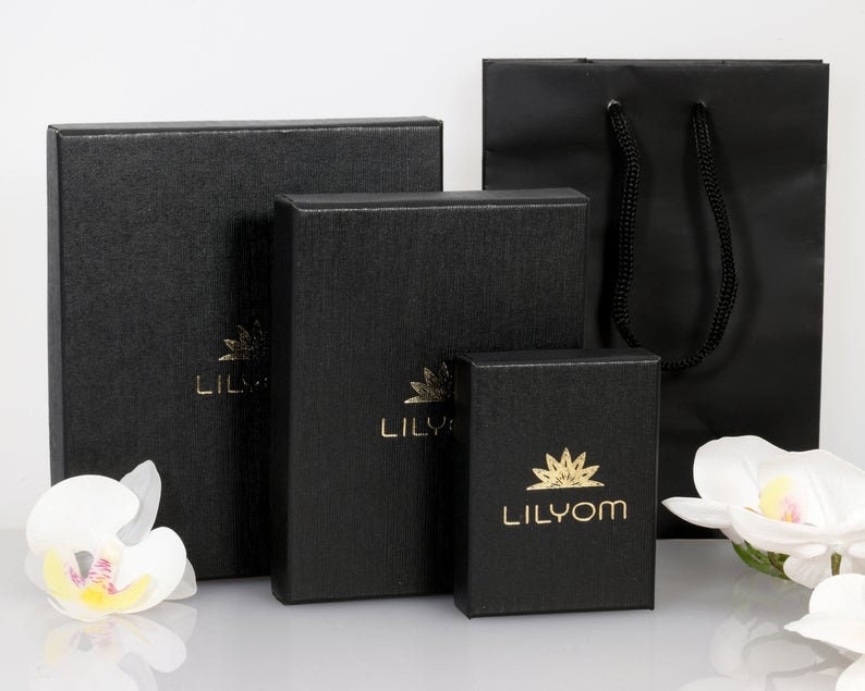 May include: Three black gift boxes with gold lettering. The smallest box has the text "Lilyom" on it. The middle box has the text "Lily" on it. The largest box has the text "Lily" on it. The boxes are sitting on a white surface.