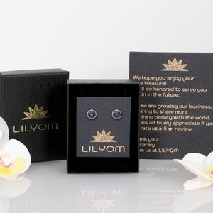 May include: A pair of small, round, blue earrings with a gold setting. The earrings are displayed in a black box with a gold logo that reads "LILYOM".