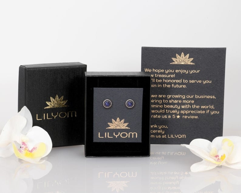 May include: A pair of small, round, blue earrings with a gold setting. The earrings are displayed in a black box with a gold logo that reads "LILYOM".