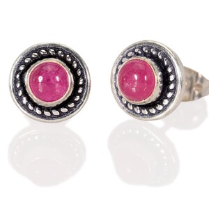 May include: A pair of silver stud earrings with a pink gemstone set in a circular bezel. The earrings have a decorative border around the gemstone.