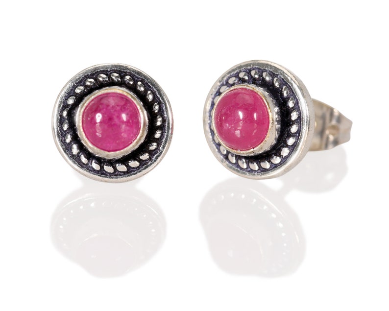 May include: A pair of silver stud earrings with a pink gemstone set in a circular bezel. The earrings have a decorative border around the gemstone.