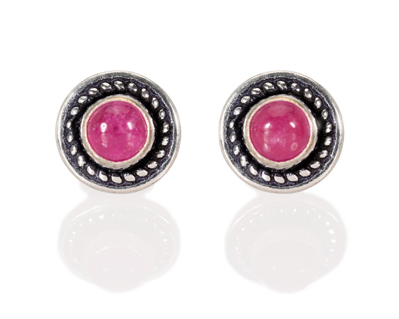 May include: A pair of silver stud earrings with a pink gemstone set in a silver bezel. The earrings have a decorative border.