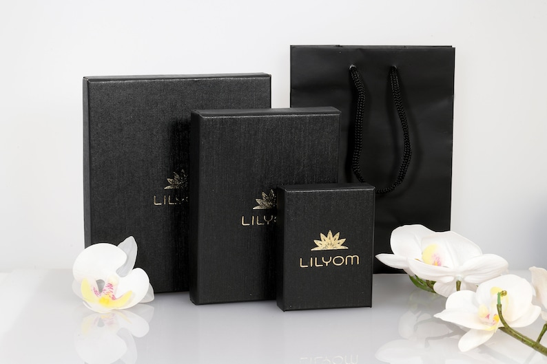 May include: Three black gift boxes with gold lettering, the smallest box says "Lilyom", the middle box says "Lily", and the largest box says "Lily". The boxes are sitting on a white surface in front of a black shopping bag with black handles.