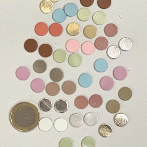 Peut inclure: Un assortiment de petites pièces rondes en cuir de différentes couleurs, dont or, rose, bleu et marron. Une pièce d'un euro est incluse pour l'échelle. Les pièces sont disposées sur une surface claire.