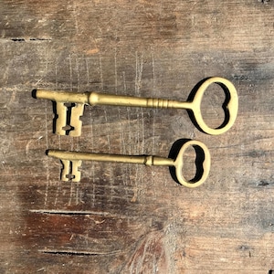 May include: Two antique brass keys, one large and one small, lie on a weathered wooden surface. The keys have ornate heart-shaped keyholes and detailed shafts. The wood grain and knots add to the vintage aesthetic.