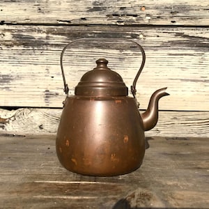 Puede incluir: Una tetera de color cobre con un cuerpo redondeado, un pico curvo y un asa. La tetera tiene una tapa y está sobre una superficie de madera. El fondo es una pared de madera desgastada. La tetera probablemente se usa para hervir agua.