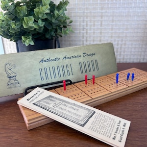 May include: A wooden cribbage board with red and blue pegs, accompanied by a vintage instruction card labeled Drueke's Cribbage Master and a box lid featuring the text Authentic American Design Cribbage Board and a knight chess piece illustration.