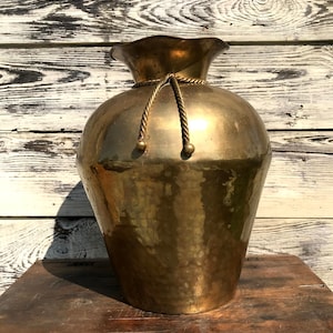 Puede incluir: Un jarrón grande de latón martillado con un cuerpo ancho y redondeado y un borde con volantes. Un detalle decorativo de cuerda con pequeñas esferas de latón adorna el cuello. El jarrón se encuentra sobre una superficie de madera envejecida.
