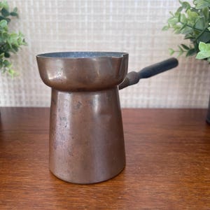 May include: A copper coffee pot with a wide, flared rim and a small pouring spout. The pot features a cylindrical body with a dark, weathered patina and a single, straight handle extending from the side, finished with a dark, matte wooden grip.