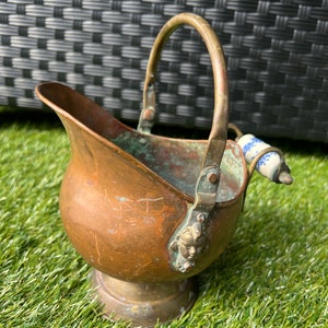 May include: A copper coal scuttle with a handle and a porcelain knob. The scuttle has a cherub face on the side and is sitting on a green surface.