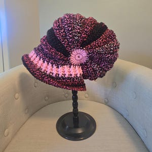 May include: A handmade crochet hat in shades of pink, black, and white, displayed on a black stand. The hat features a brim with a pink and white pattern, a black band, and a pink flower accent. The hat is a unique, handcrafted accessory.