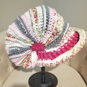May include: A crocheted hat in shades of pink, white, gray, and green. The hat is shaped like a seashell and is displayed on a black stand.