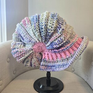 May include: A crocheted hat in shades of pink, purple, and green, shaped like a seashell, displayed on a black stand.