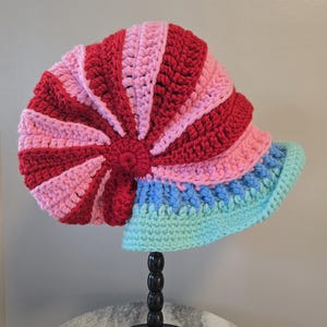 May include: A handmade crochet hat in a spiral design. The hat features alternating sections of red and pink, transitioning to blue and aqua at the brim. The hat is displayed on a black stand.