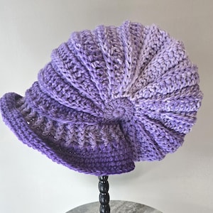 May include: A handmade, purple crochet hat with a brim. The hat features a gradient of purple hues, transitioning from a deep violet to a lighter lavender. The textured stitch work creates a ribbed effect, and the hat is displayed on a black stand.