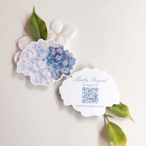 May include: A die-cut response card featuring a watercolor illustration of blue and purple hydrangea flowers. The card includes the text Kindly Respond by August 3rd, a blue QR code, and the website address WEDDINGWEBSITE.COM in blue font.