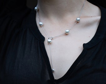 Könnte beinhalten: Eine zarte Silberkette mit sechs runden, weißen Perlen. Die Halskette hat ein Y-förmiges Design mit einer einzelnen Perle am unteren Ende des Y. Die Halskette wird über einem schwarzen Oberteil getragen.